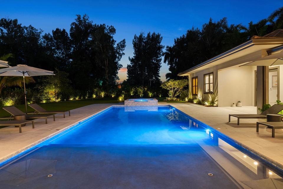 Lit pool and spa at dusk beside a single-storey villa and tall trees, Villa Banyan in Sunny Isles Beach