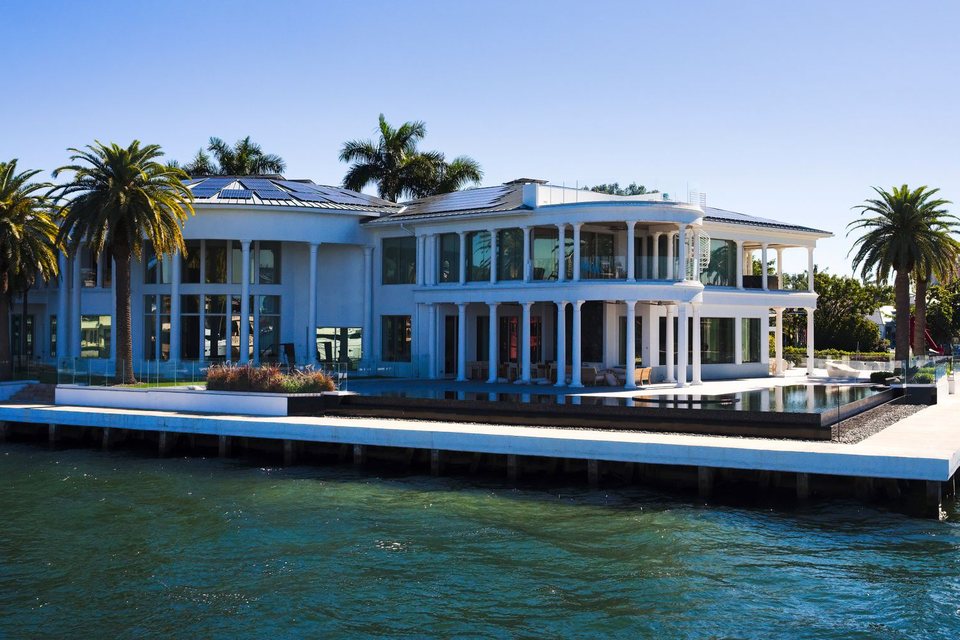 White waterfront mansion with palm trees and a private dock on turquoise bay water