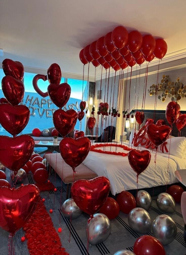 Hotel room styled for an anniversary with red heart balloons, rose petals and a love balloon on the bed