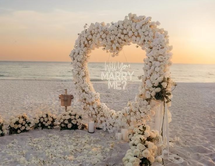 Beach proposal setup with a white rose heart arch and a Will You Marry Me neon sign at sunset