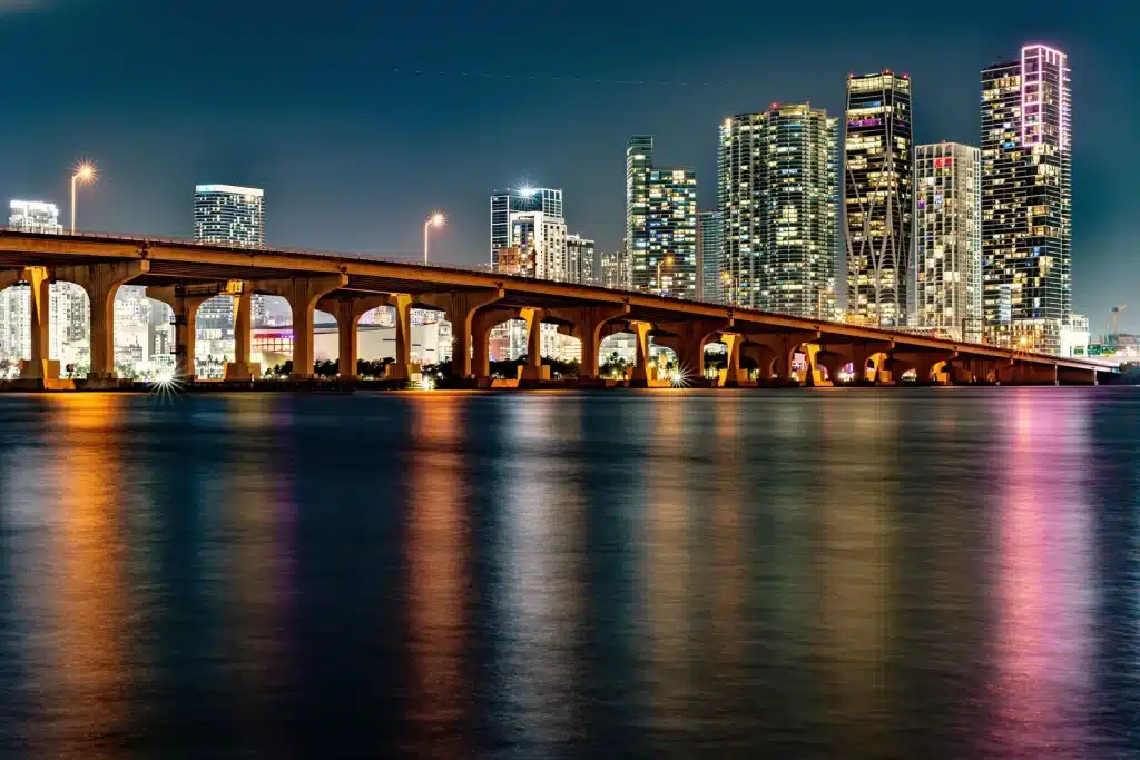 Downtown Miami towers lit at night behind a causeway, reflected in the calm water of Biscayne Bay